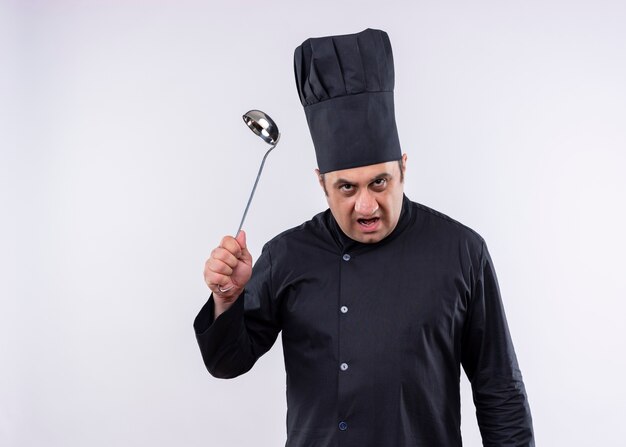 Cuoco unico maschio che indossa l'uniforme nera e cappello da cuoco che tiene il mestolo che guarda l'obbiettivo con la faccia arrabbiata che sta sopra fondo bianco