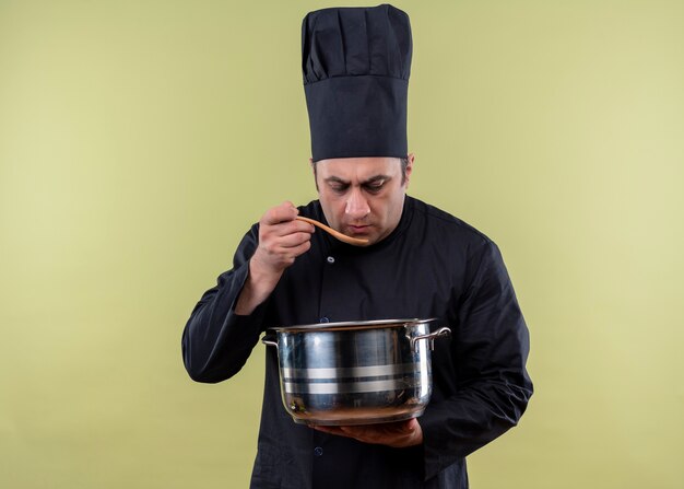 Cuoco unico maschio che indossa l'uniforme nera e cappello da cuoco che tiene la casseruola che assaggia il cibo con un cucchiaio di legno che sta sopra fondo verde