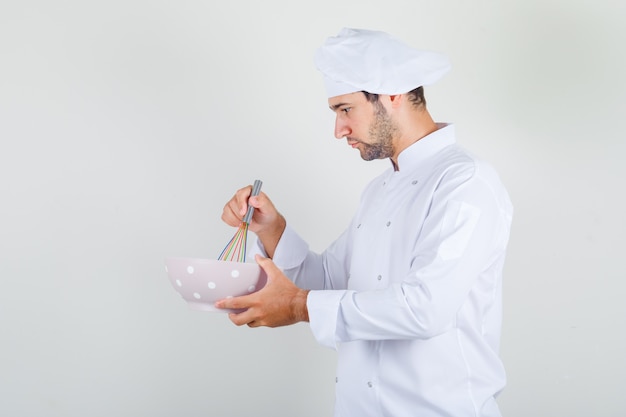 Cuoco unico maschio che mescola gli ingredienti con la frusta in una ciotola in uniforme bianca e sembra occupato