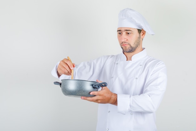 Cuoco unico maschio che mescola la minestra con il cucchiaio di legno in uniforme bianca