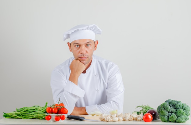 Cuoco unico maschio che si siede e che esamina macchina fotografica in uniforme e cappello in cucina