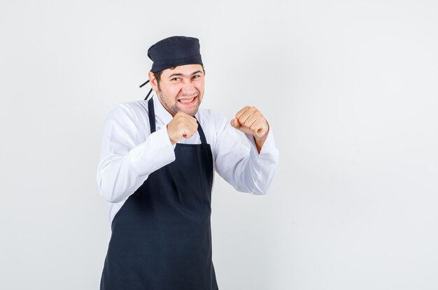 Cuoco unico maschio che stringe i denti in boxer posa in uniforme, grembiule e sembra allegro. vista frontale.