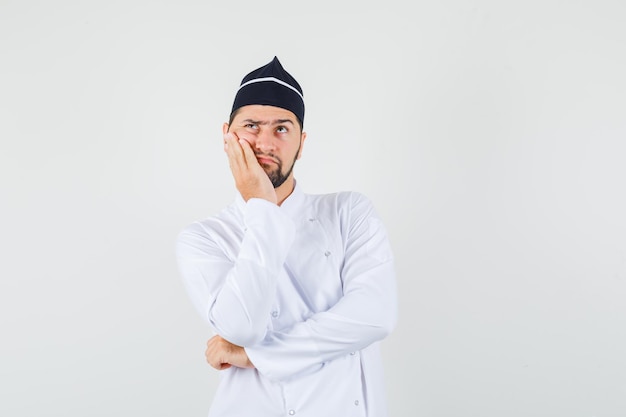 Cuoco unico maschio che tiene il palmo sulla guancia in uniforme bianca e sembra pensieroso. vista frontale.