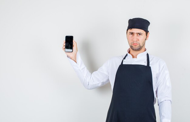 Cuoco unico maschio che tiene il telefono cellulare in uniforme, grembiule e guardando sconvolto, vista frontale.