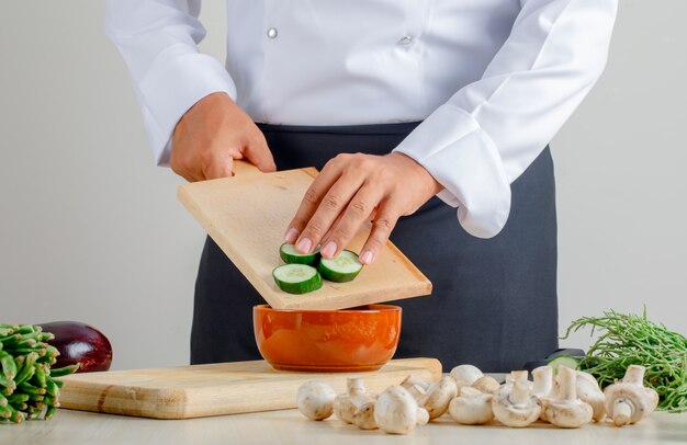 Cuoco unico maschio che versa cetriolo affettato nella ciotola in cucina in uniforme e grembiule