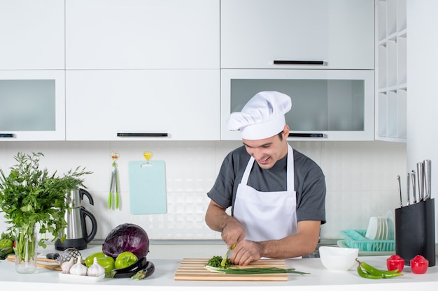 Cuoco unico maschio di vista frontale in uniforme che taglia i verdi dietro il tavolo della cucina