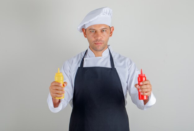 Cuoco unico maschio in cappello, grembiule e divisa in possesso di bottiglie di ketchup e senape