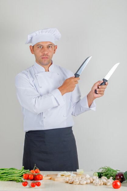 Cuoco unico maschio in uniforme, cappello e grembiule che tengono i coltelli metallici in cucina