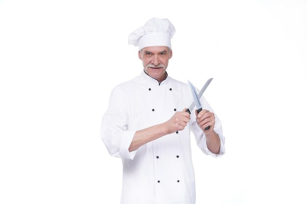Cuoco unico maschio in uniforme che tiene due coltelli di metallo mentre rimane sul muro bianco