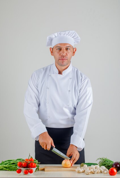 Cuoco unico maschio in uniforme, grembiule e cappello che tagliano cipolla sul bordo di legno in cucina