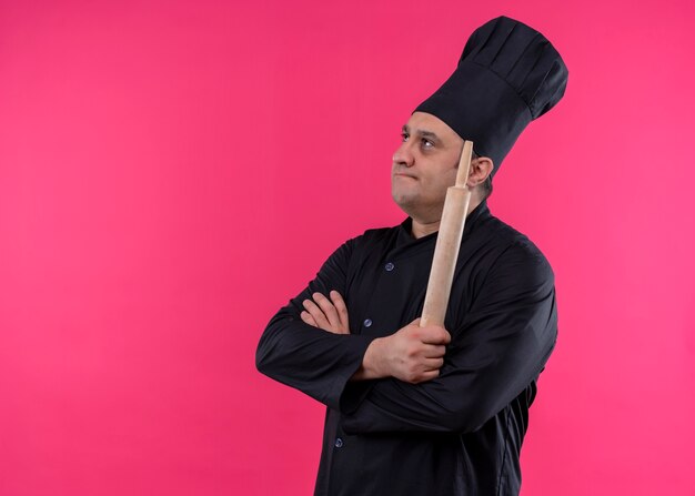 Cuoco unico maschio premuroso che indossa l'uniforme nera e cappello da cuoco che tiene il mattarello che guarda da parte con espressione pensierosa in piedi su sfondo rosa