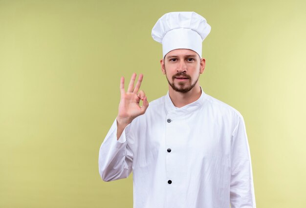 Cuoco unico maschio professionista cuoco in uniforme bianca e cappello da cuoco che mostra gesto giusto che sembra fiducioso in piedi su sfondo verde