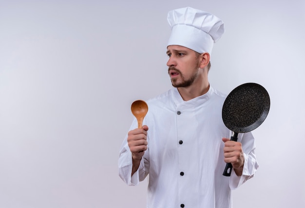 Cuoco unico maschio professionista cuoco in uniforme bianca e cappello da cuoco che tiene padella e cucchiaio di legno che sembra preoccupato in piedi su sfondo bianco