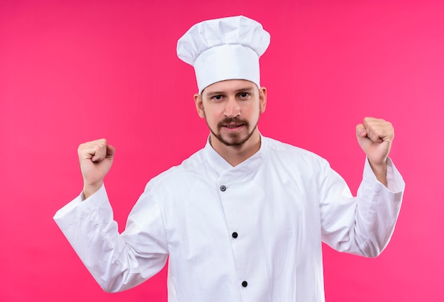 Cuoco unico maschio professionista cuoco in uniforme bianca e cappello da cuoco stringendo i pugni felici ed emozionati rallegrandosi del suo successo in piedi su sfondo rosa