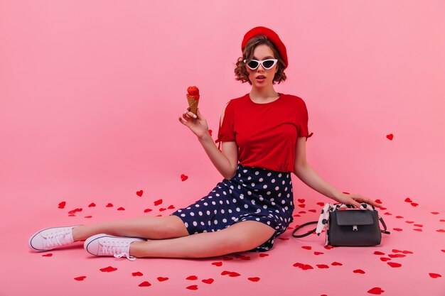 Curiosa giovane donna in gonna tenendo il gelato. fiduciosa ragazza francese in berretto che mangia dessert.