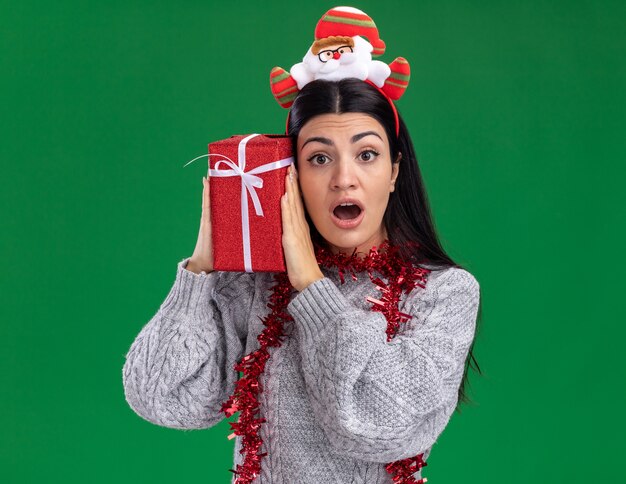 Curiosa giovane ragazza caucasica indossando la fascia di Babbo Natale e la ghirlanda di orpelli intorno al collo tenendo il pacchetto regalo toccando la testa con esso guardando la telecamera isolata su sfondo verde