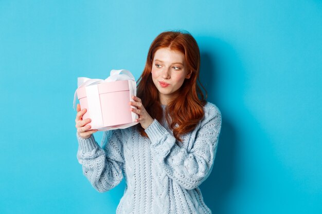 Curiosa ragazza adolescente con i capelli rossi, scuotendo la confezione regalo e chiedendosi cosa c'è dentro, in piedi su sfondo blu