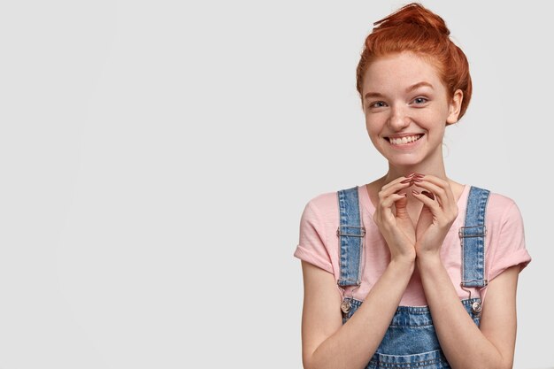 Curiosa ragazza adolescente felice con la pelle lentigginosa, capelli rossi, tiene le mani unite, guarda con espressione astuta, aspetta aiuto, vestita in tuta, isolato su muro bianco, spazio libero da parte