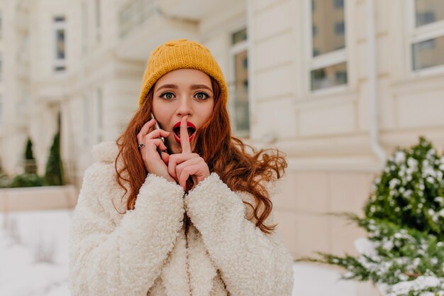 Curiosa ragazza dai capelli rossi in cappello parlando al telefono all'aperto. Donna abbastanza giovane che chiama qualcuno.