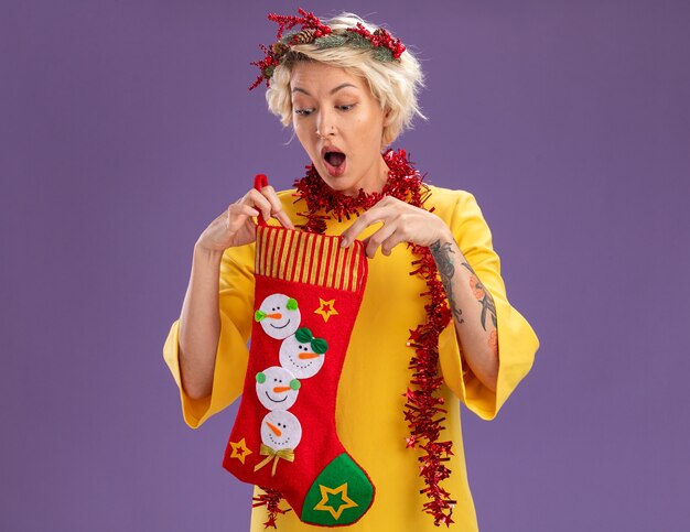 Curioso giovane donna bionda che indossa la testa di Natale ghirlanda e orpello ghirlanda intorno al collo tenendo la calza di Natale guardando al suo interno isolato su sfondo viola