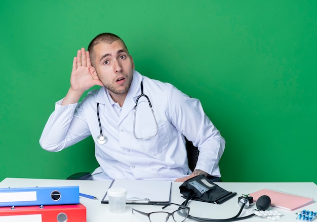 Curioso giovane medico maschio che indossa abito medico e stetoscopio seduto alla scrivania con strumenti di lavoro facendo non può sentirti gesto isolato sul verde