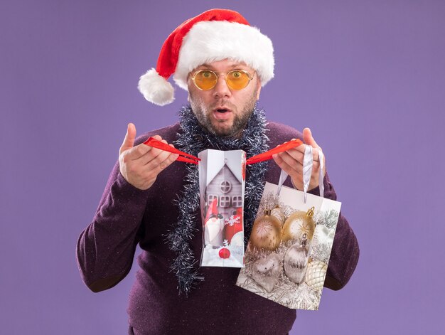 Curioso uomo di mezza età che indossa un cappello da Babbo Natale e una ghirlanda di orpelli intorno al collo con gli occhiali che tengono i sacchetti del regalo di Natale che aprono uno che guarda l'obbiettivo isolato su fondo viola