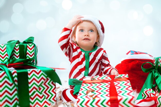 Cute baby girl 1 anno di età che indossa il cappello della santa in posa su decorazioni natalizie con doni. Seduto sul pavimento con la palla di Natale