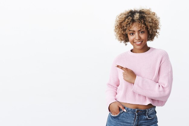 Dai un'occhiata con me. Gioiosa ed elegante donna attraente sicura di sé con acconciatura afro bionda in maglione caldo invernale che punta all'angolo in alto a sinistra e sorride come invitante o promuovendo lo spazio della copia