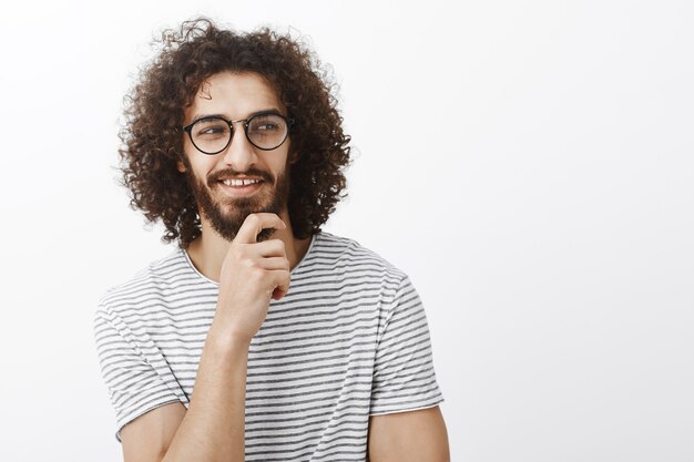 Dai un'occhiata. Ragazzo affascinante e innamorato incuriosito in uno stato d'animo romantico giocoso, tenendo la mano sulla barba, guardando a destra e sorridendo con un'espressione curiosa e compiaciuta