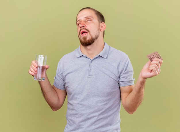 Debole giovane bello slavo malato che tiene il pacchetto di capsule e bicchiere d'acqua con gli occhi chiusi isolati sulla parete verde oliva