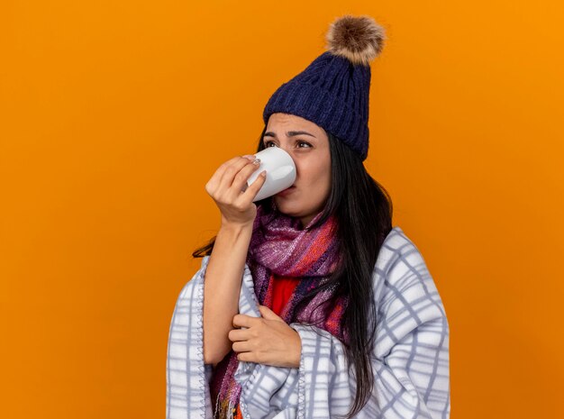 Debole giovane donna malata che indossa cappello invernale e sciarpa avvolta in un plaid in piedi in vista di profilo che beve tazza di tè guardando dritto isolato sul muro arancione