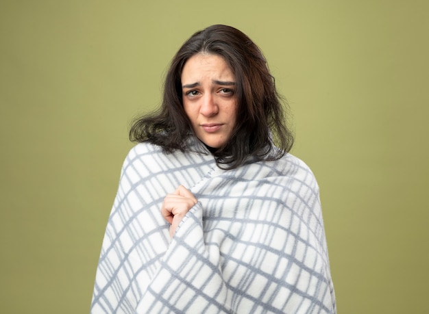 Debole giovane indoeuropeo ragazza malata che indossa accappatoio avvolto in plaid guardando la telecamera afferrando plaid isolato su sfondo verde oliva con spazio di copia