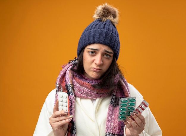 Debole giovane ragazza malata caucasica che indossa un abito invernale cappello e sciarpa con confezioni di pillole mediche che guarda l'obbiettivo isolato su sfondo arancione