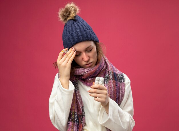 Debole giovane ragazza malata con gli occhi chiusi che indossa una veste bianca e cappello invernale con sciarpa che tiene pillole e siringa mettendo la mano sulla fronte isolata sul rosa