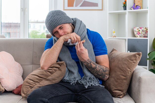 Debole giovane uomo malato che indossa sciarpa e cappello invernale seduto sul divano in soggiorno tenendo un pacchetto di capsule e tovagliolo asciugandosi il naso con la mano con gli occhi chiusi