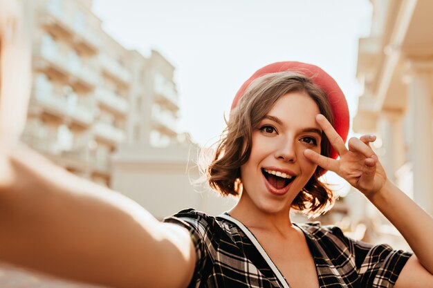 Debonair donna in berretto francese in posa sulla strada con il segno di pace. Foto all'aperto di una ragazza affascinante con gli occhi scuri.