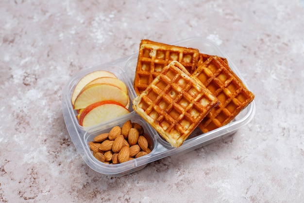 Deliziosa colazione con mandorle, fette di mele rosse, waffle, cornetti su scatola di plastica per il pranzo alla luce