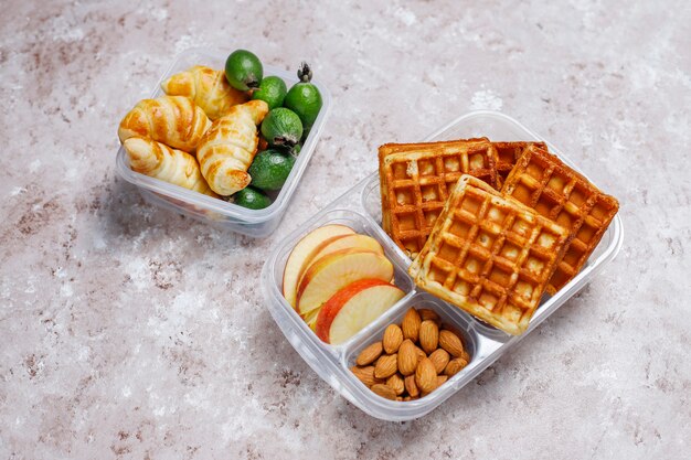 Deliziosa colazione con mandorle, fette di mele rosse, waffle, cornetti su scatola di plastica per il pranzo alla luce