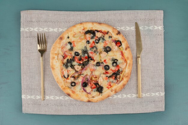 Deliziosa pizza calda con olive e pomodori sul tavolo blu con posate d'oro.