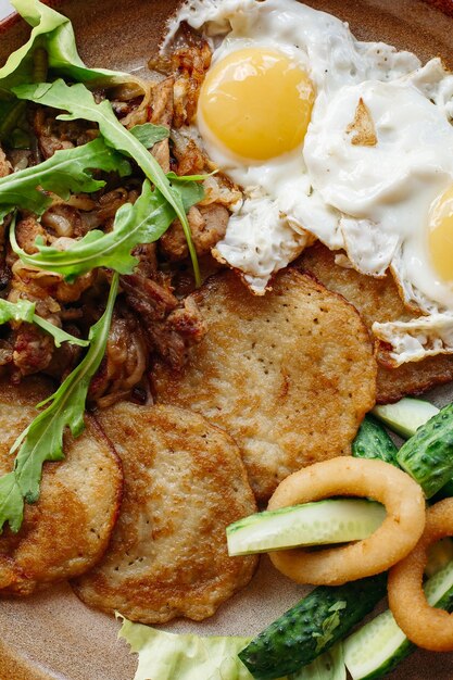 Deliziosi pancake di patate sdraiati su un piatto di argilla marrone con uova fritte Cetrioli verdi freschi cipolla dorata fritta e carne croccante serviti con foglie di insalata Gustoso pasto mattutino appetitoso