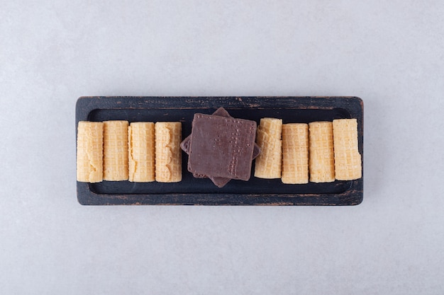 Deliziosi rotoli di wafer e wafer ricoperti di cioccolato su un piatto di legno sul marmo.