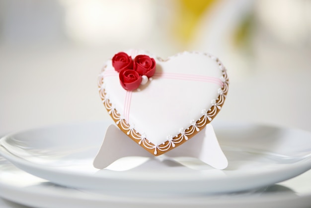 Delizioso biscotto di pan di zenzero ricoperto di glassa bianca dolce e decorato con roselline rosse e piccole perle bianche si trova sul tavolo con piatto bianco. Buona decorazione per la tavola nuziale festiva.