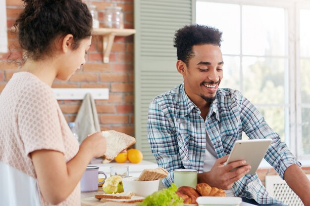 Delizioso giovane con la pelle scura e i capelli ricci legge ad alta voce storie divertenti sul tablet PC mentre siede di fronte alla moglie che è occupata