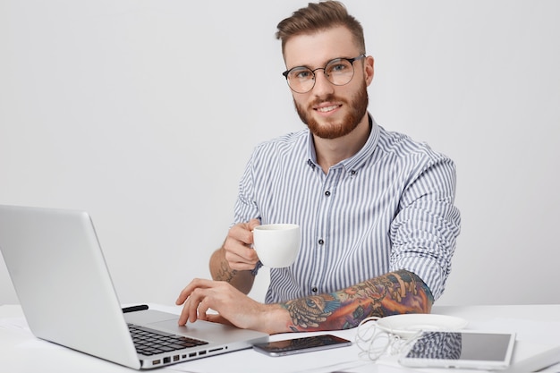 Delizioso libero professionista con una folta barba, indossa occhiali, ha tatuaggi, lavora a distanza