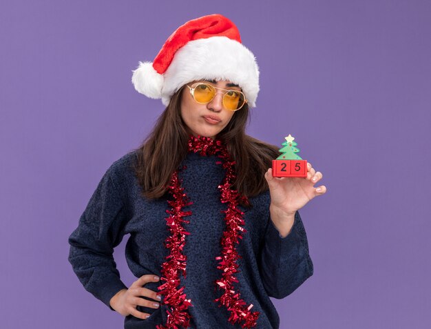 Delusa giovane ragazza caucasica in occhiali da sole con cappello da Babbo Natale e ghirlanda intorno al collo tiene l'ornamento dell'albero di natale isolato sulla parete viola con spazio di copia