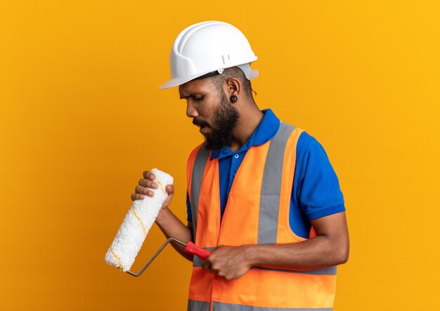Deluso giovane costruttore afro-americano uomo in uniforme con casco di sicurezza che tiene e guardando il rullo di vernice isolato su sfondo arancione con spazio di copia