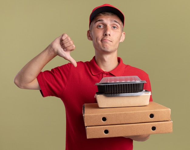 Deluso giovane ragazzo delle consegne biondo pollice in giù e tiene i pacchetti di cibo su scatole per pizza isolate su parete verde oliva con spazio di copia