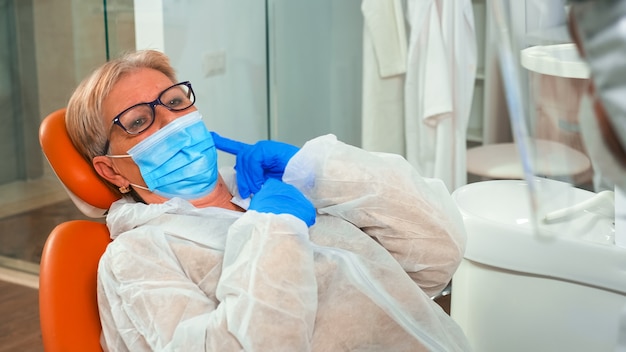 Dentista e donna con maschera di protezione che parlano di problemi dentali in clinica stomatologica durante la pandemia globale. Medico con visiera, guanti, equipaggiamento protettivo che esamina paziente anziano