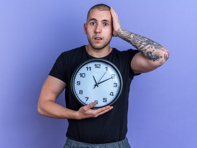 Deplorato giovane bel ragazzo che indossa una maglietta nera che tiene l'orologio da parete che mette la mano sulla testa isolata sul muro blu