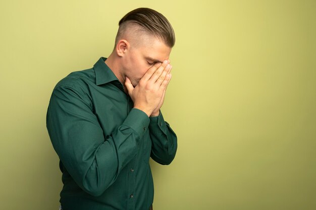 Deprimente giovane uomo bello in camicia verde che copre il viso con le braccia stressate e nervose